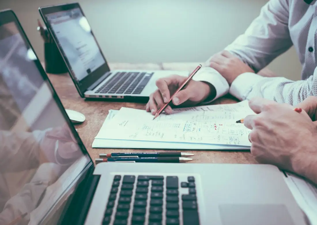 People marking printed plans beside open laptops on a desk